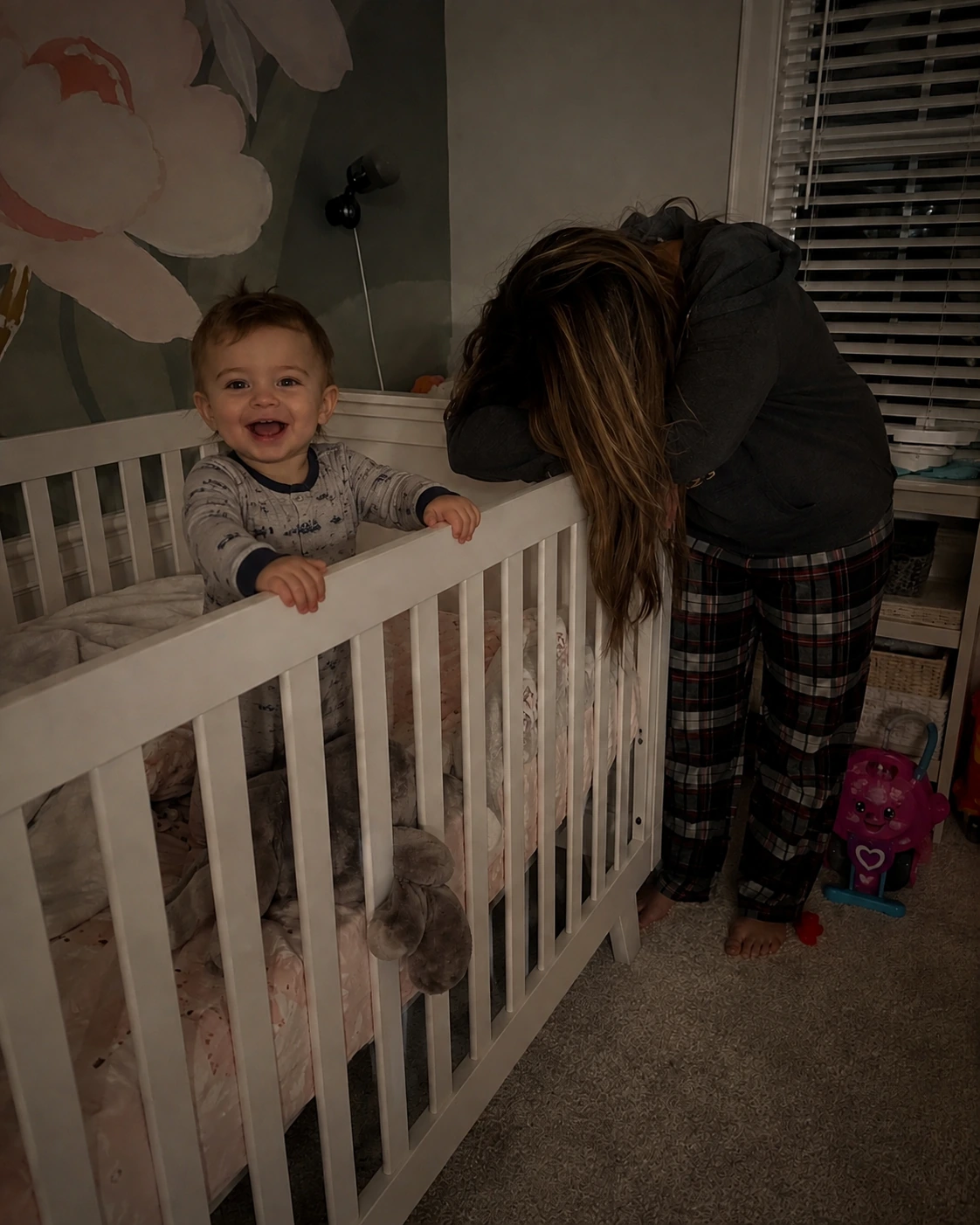 Happy toddler standing in crib while exhausted mother rests her head on the railing